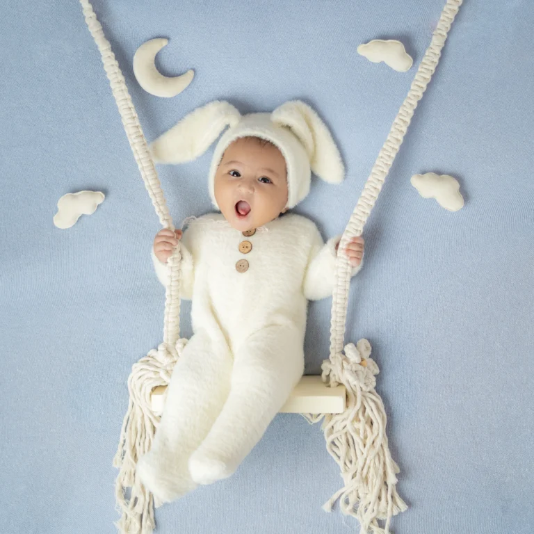 Newborn dressed in a fluffy bunny outfit with playful ears, enjoying a swing set against a dreamy sky backdrop.